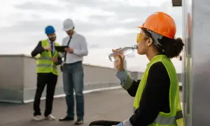 Kobieta w kasku i kamizelce odblaskowej pije wodę, odpoczywając od pracy w wysokich temperaturach. Dwóch mężczyzn w tle dyskutuje nad tabletem.