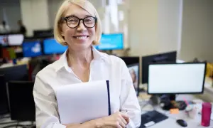 Uśmiechnięta kobieta z blond włosami i okularami, trzymająca teczkę. Jej emerytura z Anglii w Polsce przebiega pomyślnie.