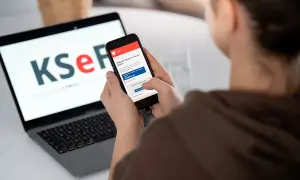 Kobieta korzysta z telefonu, na którym widnieje aplikacja KSeF. Na laptopie w tle logo KSeF. To KSeF krok po kroku.