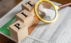 Drewniane klocki z literami "PIT" leżą na formularzu podatkowym. Złota lupa bada dokument, sugerując, jak zapłacić podatek dochodowy.