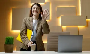 Uśmiechnięta kobieta w okularach macha ręką, trzymając teczkę i kawę. Czy dzień próbny jest płatny?