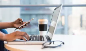 Kobieta pracuje na laptopie, trzymając telefon. Obok stoi kubek kawy. To idealne miejsce do planowania strategii rozwoju firmy, być może z uwzględnieniem wsparcia pomoc de minimis.