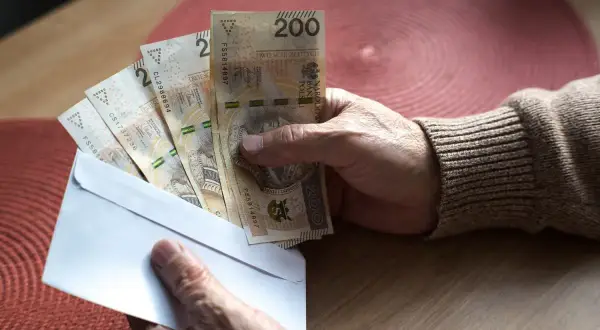 Dłoń trzyma kopertę z banknotami 200 zł. Może to być związane z nowymi zasadami przejścia na emeryturę, tabela z wyliczeniami jest gdzieś obok.
