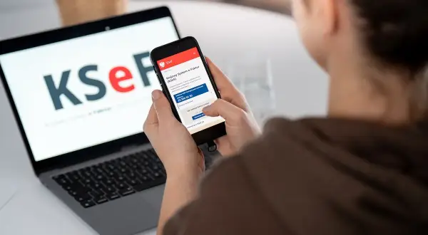 Kobieta korzysta z telefonu, na którym widnieje aplikacja KSeF. Na laptopie w tle logo KSeF. To KSeF krok po kroku.