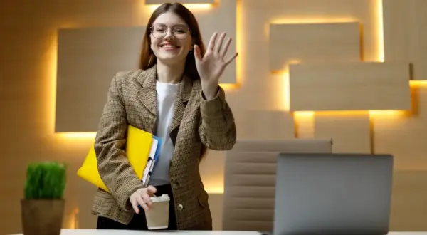 Uśmiechnięta kobieta w okularach macha ręką, trzymając teczkę i kawę. Czy dzień próbny jest płatny?