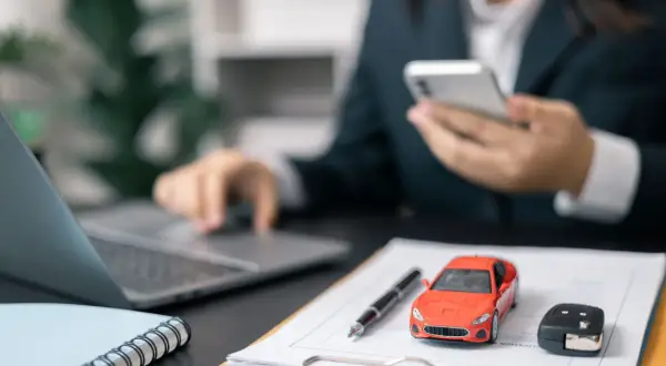 Biznesmen analizuje amortyzację samochodu osobowego 75 czy 100, korzystając z telefonu i laptopa. Na biurku model auta i kluczyki.