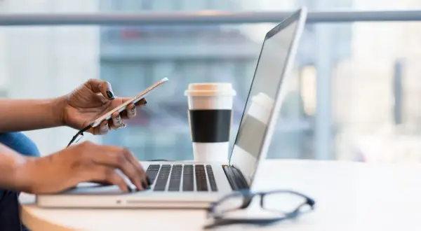 Kobieta pracuje na laptopie, trzymając telefon. Obok stoi kubek kawy. To idealne miejsce do planowania strategii rozwoju firmy, być może z uwzględnieniem wsparcia pomoc de minimis.