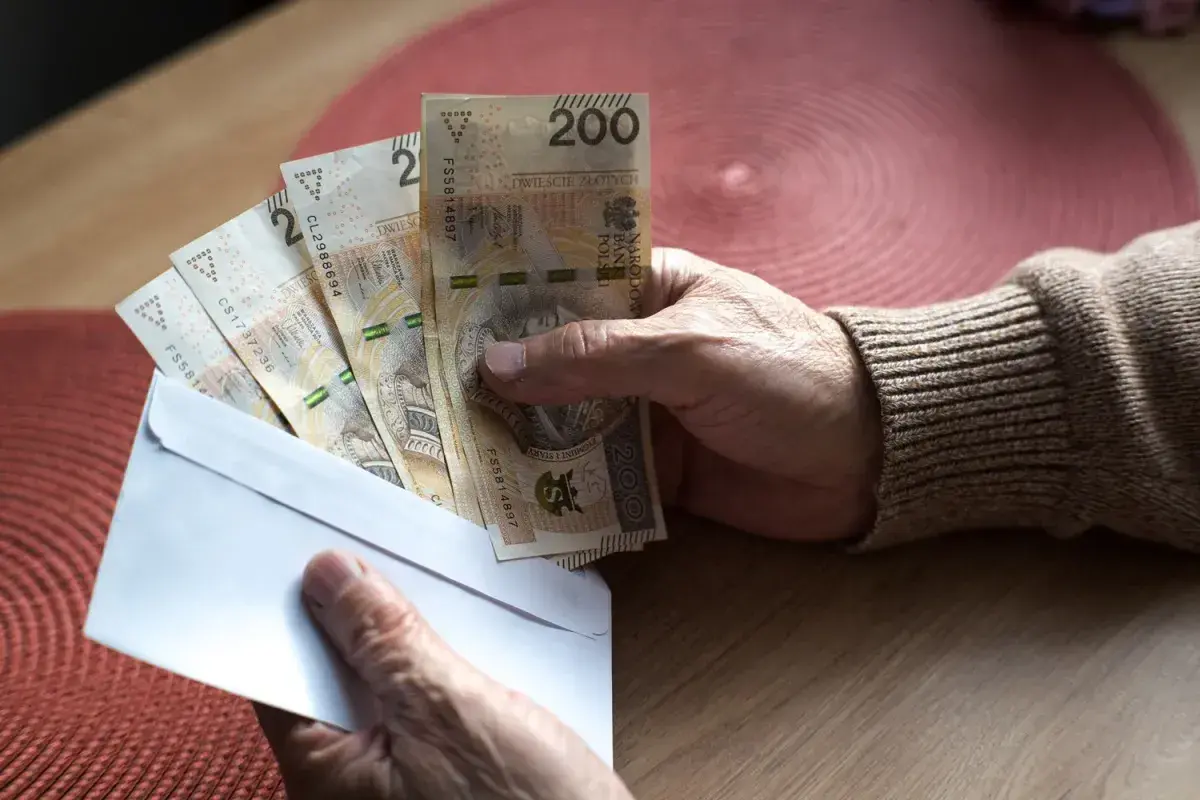 Dłoń trzyma kopertę z banknotami 200 zł. Może to być związane z nowymi zasadami przejścia na emeryturę, tabela z wyliczeniami jest gdzieś obok.