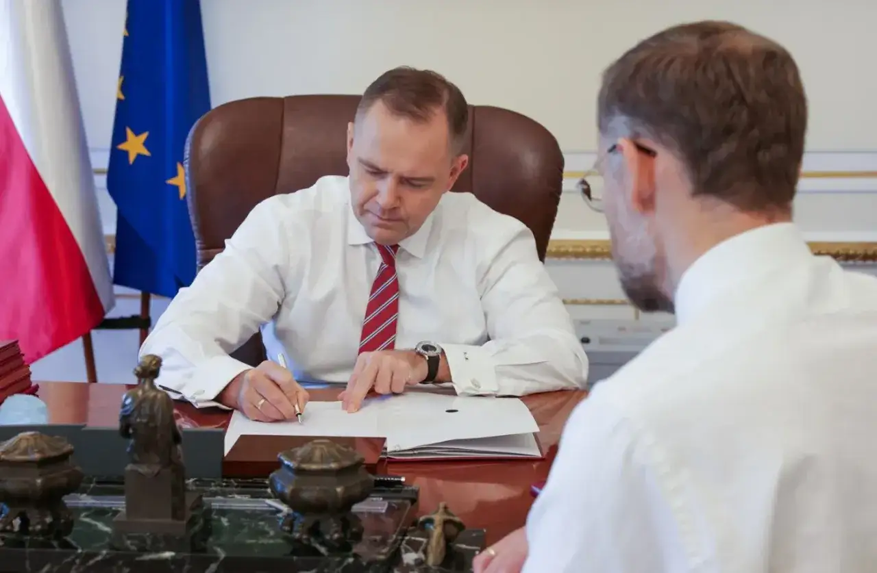 Mężczyzna w białej koszuli i krawacie podpisuje umowę o dzieło przy biurku, obok flagi Polski i UE.