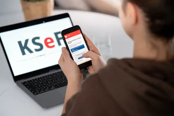Kobieta korzysta z telefonu, na którym widnieje aplikacja KSeF. Na laptopie w tle logo KSeF. To KSeF krok po kroku.