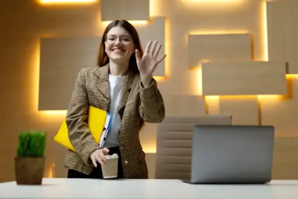 Uśmiechnięta kobieta w okularach macha ręką, trzymając teczkę i kawę. Czy dzień próbny jest płatny?