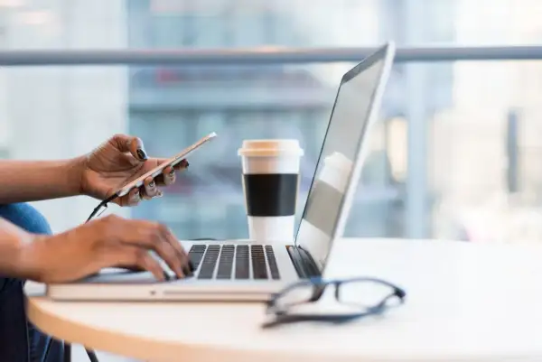 Kobieta pracuje na laptopie, trzymając telefon. Obok stoi kubek kawy. To idealne miejsce do planowania strategii rozwoju firmy, być może z uwzględnieniem wsparcia pomoc de minimis.
